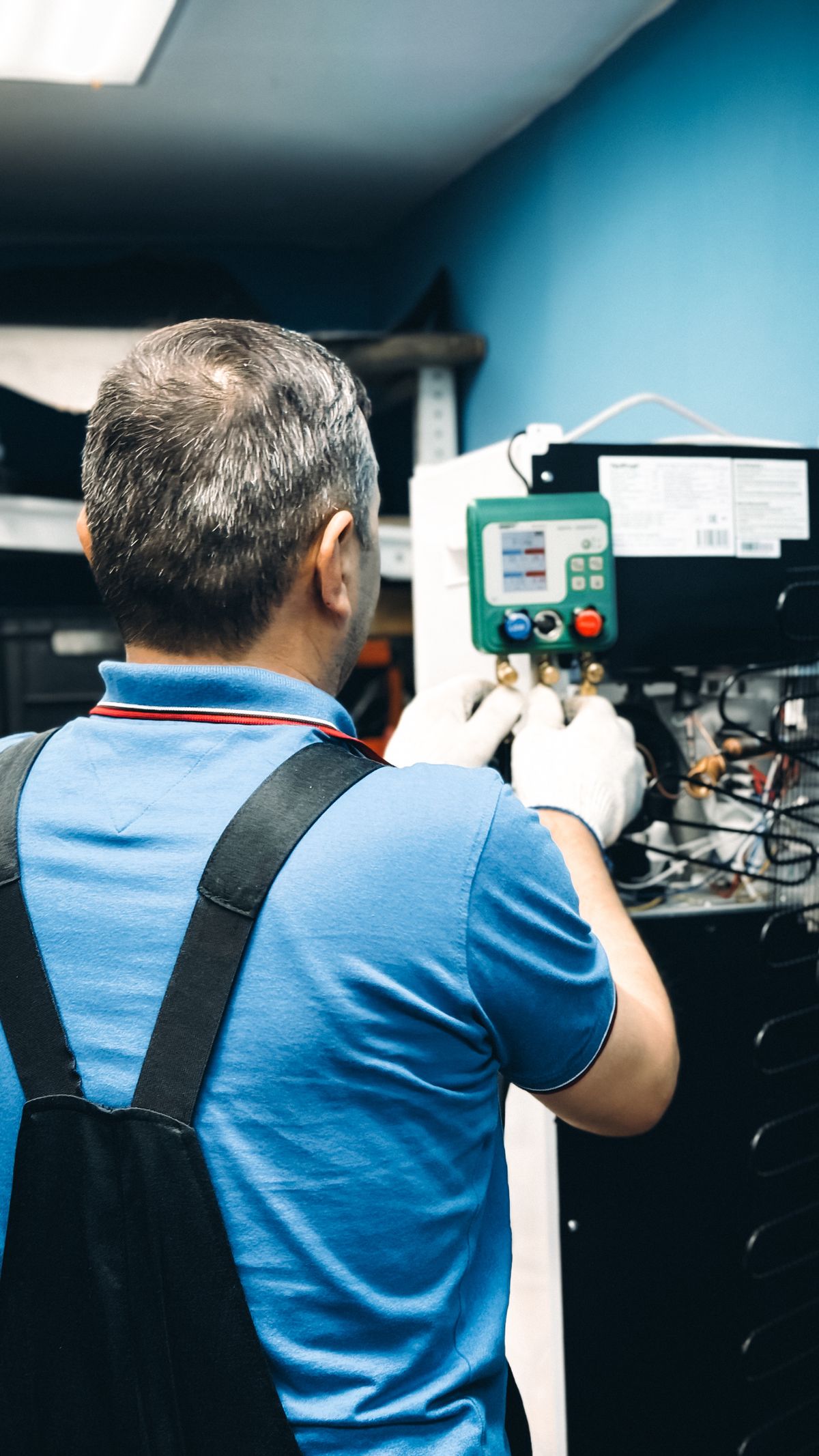 HVAC technician in a blue uniform using refrigerant manifold gauges to service an air-handling unit indoors