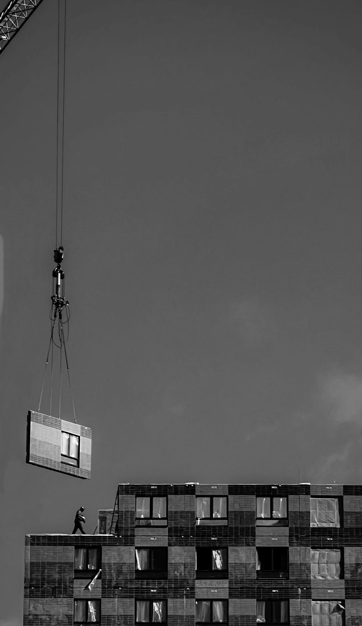 Construction crane hoisting a large prefabricated unit onto the top of a building while a worker walks the roof edge